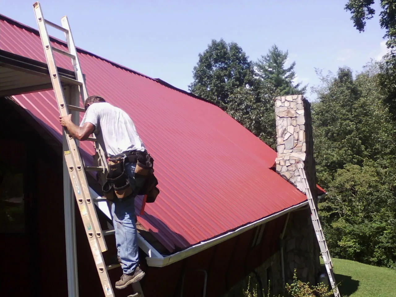 Roofer on a ladder beside a red metal roof and stone chimney — Soffit & Fascia Repair by Roofing Brothers in Big Run